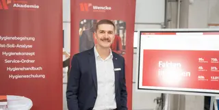 Ein Mann mit braunen Haaren und Schnauzer in blauem Anzug und weißem Hemd, der vor zwei roten Rollups der Wencke Akademie steht